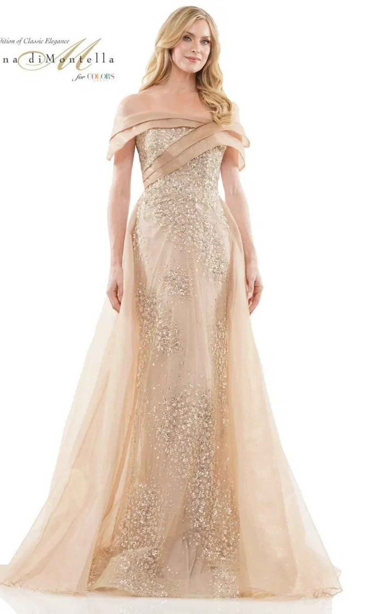A woman models the Rina Di Montella RD2816 long formal dress in champagne, featuring an off-the-shoulder design, floor-length tulle overskirt, sheer glitter overlay, and set against a plain background.